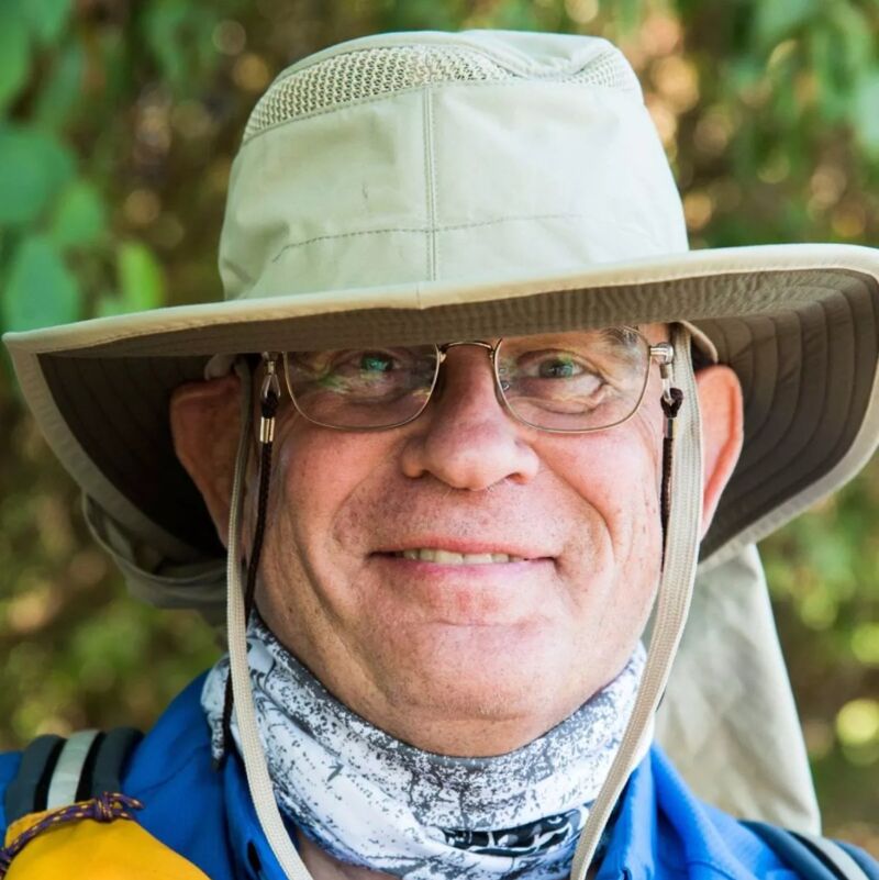 The image shows a close-up of an older man wearing glasses, a hat, and a neck gaiter. He is smiling at the camera. The background is blurred, suggesting an outdoor setting, possibly a park or a trail. The man appears to be enjoying the outdoors.